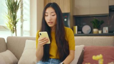 Güzel Asyalı genç bayan evdeki kanepede otururken telefon kullanıyor. Sosyal medya mesajları uzak iletişimin tadını çıkarıyor. Kapatın. Yavaş çekim