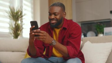 Mutlu ve genç Afrikalı Amerikalı adam telefon kullanıyor gülümsüyor iyi haberler okuyor evdeki kanepede oturuyor. Sosyal medyada iletişim kurmak iş uygulamasını gevşetiyor. Yakışıklı adammış. - İnternet. Yavaş çekim