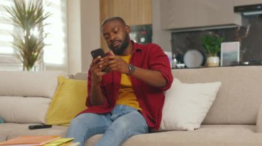 Çekici, Afro-Amerikalı bir adam telefonu kullanıyor. Telefonu masaya koyuyor. Evdeki kanepede oturuyor. Sosyal medyada iletişim kurmak iş uygulamasını gevşetiyor. Yakışıklı adammış. - İnternet. Yavaş çekim