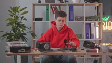 Kırmızı kapüşonlu genç, sevimli, konsantre öğrencinin uzaktan çekilen fotoğrafı. Modern işyerinde kuadkopteri sıkıştıran zeki Ukraynalı erkek mühendis. Modern teknoloji, alet. FPV teknolojisi. Eğitim. Bilimsel kavram.