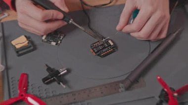 Kırmızı spor elbiseli erkeklerin yakın çekim görüntüleri masada oturup bordaya lehimleme yapıyorlar. Mühendis lehim kullanıyor. Sıradan bir iş günü. Modern teknoloji. Drone prototipi. Hava konsepti.