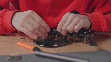 Kırmızı kapüşonlu genç adamın yakın çekim görüntüsü Quadcopter 'ın detaylarını onarıyor. Beyazların insansız hava aracını tamir ederken çekilmiş. İnsansız hava aracı. Modellik. Ekipman parçaları. Kaliteli iş. Hava robotik mekanizması.