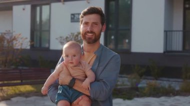 Küçük bebeği kucağına alıp doğrudan kameraya bakarken mutlu bir babanın portresi. Modern binaların arka planında güzel görünüşlü, gülümseyen beyaz bir adamın güzel bir oğlu var. Aile