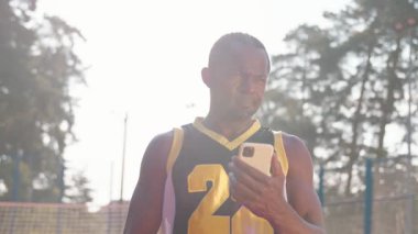 Arkadaş canlısı görünümlü düşünceli Afrikalı basketbolcu elinde cep telefonu tutarak sohbet ediyor. Atletik gülümseyen adam akıllı telefon ekranına bakıyor, mesaj atıyor. Dışarısı, faaliyet, mahkeme. Modern teknoloji
