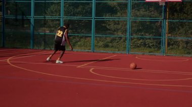 Spor kıyafetleri içinde yakışıklı Afro-Amerikan bir adamın şehir spor sahasında basketbol oynaması. Çekici, profesyonel bir basketbolcunun portresi. Egzersiz yap. Sabah vakti, yaz.