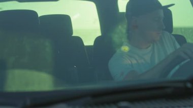 Arabasında oturan, günlük giysiler içinde beyaz bir adamın yakın plan fotoğrafı. Adam kapıyı açıyor ve arabadan iniyor. Saha. Kasaba dışında bir köy. Açık hava.