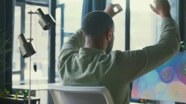 Ofiste çalışan ve elleri arkada oturan bir ofis çalışanının arka görüntüsü. Günlük giysiler içinde kaygısız Afrikalı Amerikalı bir adam. İçeride. Başarı, profesyonelce. Teknoloji. İşte mola.