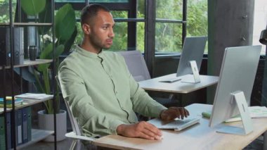 Modern ofisteki işyerinde oturan, klavyede fare kullanarak daktilo yazan çekici, genç ve odaklanmış bir Afrikalı adam. Ofis çalışanı kameraya onay jesti gösteriyor. Başparmaklar yukarı.