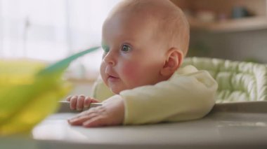 Mutfağın bulanık arka planına bakarak karnını doyurmak için bebek koltuğunda oturan meraklı bir çocuğu vurmak. Sabah oldu. İçeride. - Eve. Gündüz