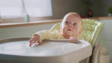 Bebek koltuğunda oturan sevimli çocuk mutfakta öğle yemeğini bekliyor. Sevimli bir bebeğin portresi. Aile kavramı. Yemek zamanı. İçeride. Çocukluk