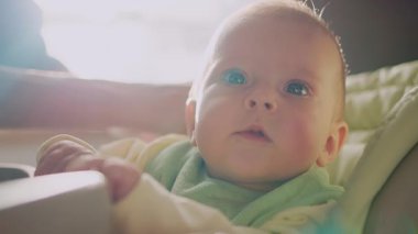 Çok fonksiyonlu bebek koltuğunda oturan küçük sevimli çocuğun yakından çekilmiş fotoğrafı ve yukarı bakıyor. Annenin elleri. Gün ışığı, gündüz. Evde, içeride.
