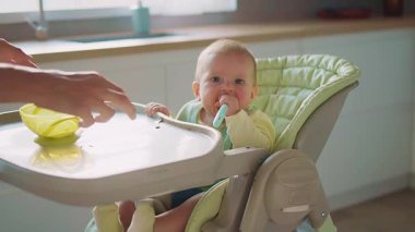 Kaşık tutan sevimli küçük erkek bebek portresi. Açık yeşil çoklu koltukta tepsiyle oturan komik, dağınık suratlı çocuk görüntüsü. İçeride. Mutfak.