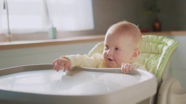 Kapalı portre, büyüleyici gülümseyen bir bebek, yüksek bir sandalyede el sallayarak etrafa bakıyor. Çocukluk. Kahvaltıda. Yavaş çekim