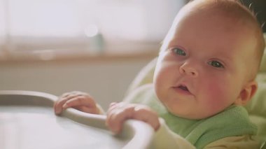 Kameraya bakarak, yüksek sandalyede oturan sevimli, gülümseyen genç bir bebek portresi. Çocukluk. Kahvaltıda. Yavaş çekim