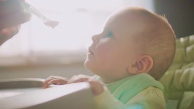 Annenin güneş ışığında küçük bebeği besleyerek yüksek sandalyeyi beslemesi ve çocuğun kameraya bakması. Yüzünde yemek lekesi olan bir çocuk. Anne yardım et. Modern apartman mutfağı. Yavaş çekim
