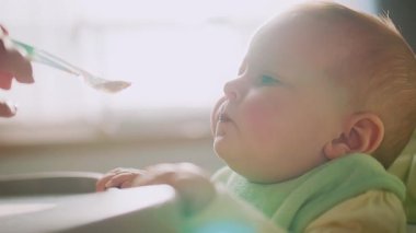Annenin küçük bebeği beslerken, sandalyeye otururken ve çocuğun kameraya bakarken ellerini kapat. Yüzünde yemek lekesi olan bir çocuk. Anne yardım et. Modern apartman mutfağı. Yavaş çekim