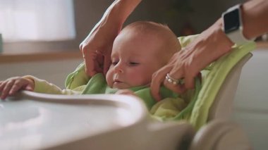 Annenin yüksek sandalyeyi beslemek için bebeği hazırlarken ellerini kapat, çocuk kendini yemeye çalışıyor. Anne yardım et. Modern apartman mutfağı. Yavaş çekim