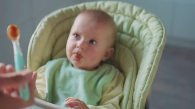 Annenin küçük bebeği yüksek sandalyeyle beslerken ellerini kapat, çocuk kendini yemeye çalışıyor. Yüzünde yemek lekesi olan bir çocuk. Anne yardım et. Modern apartman mutfağı. Yavaş çekim