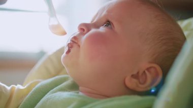 Annenin küçük bebeği yüksek sandalyeyle beslerken ellerini kapat, çocuk kendini yemeye çalışıyor. Güneş ışığı. Yüzünde yemek lekesi olan bir çocuk. Modern apartman mutfağı. Yavaş çekim