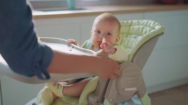 Anne, küçük bebeğini yüksek sandalyeyle beslerken kendini yemeye çalışıyor. Yüzünde yemek lekesi olan bir çocuk. Anne yardım et. Modern apartman mutfağı. Yavaş çekim