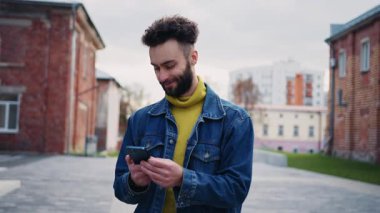 Gülümseyen genç adamın portresi sokakta yürüyor ve etrafa bakıyor. Kotlu ceketli, sarı kazaklı, akıllı telefon kullanan rahat görünümlü bir adam. Keyfin yerinde. Açık hava.