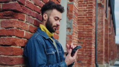 Duvarın yanında duran ve akıllı telefondan mesaj atan yakışıklı beyaz adamın yan görüntüsü. Gülümseyen çekici adamın portresi gözlerini kaçırıyor ve sokakta mesajlaşıyor. Gündüz, açık havada. Şehir