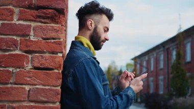 Odaklanmış, kot ceketli, akıllı telefondan mesaj atan yakışıklı bir adamın portresi. Mutlu düşünceli adam duvarın yanında dururken kızla sohbet ediyor. Binalar. Gündüz vakti. Sonbahar. Dışarıda.