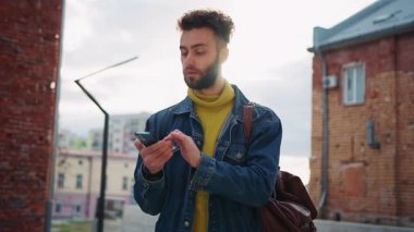 Elinde akıllı telefonuyla caddede dikilen odaklanmış genç, sakallı, beyaz bir adam görüntüsü. Alet kullanan ve evet işareti yapan yakışıklı ve başarılı bir adamın portresi. İyi haber konsepti. Dışarıda.