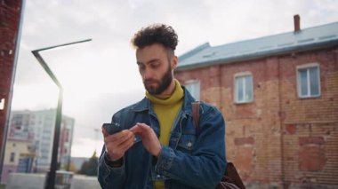 Sarı kazaklı, mavi kot ceketli, akıllı telefon ve mesaj taşıyan düşünceli ve yakışıklı genç bir adamı vurmak. Erkek hipster mesajlaşması, sosyal medya. Açık havada. Bağlantı, internet, iletişim. Şehir, yaşam tarzı