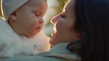 Sonbaharda parkın bulanık manzarasında annesinin elinde oturan küçük sevimli çocuğun yakın plan görüntüsü. Annem beyaz elbiseli sevimli gülümseyen bebeğe bakıyor. Annelik