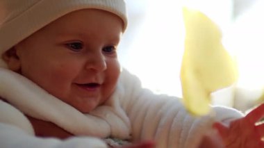 Beyaz, yumuşak elbiseli, sarı yaprağa dokunan gülümseyen küçük kızın yakın görüntüsü. Neşeli bir bebeğin portresi. Çocukluk. Açık havada. Sonbahar geldi. Güneş ışığı