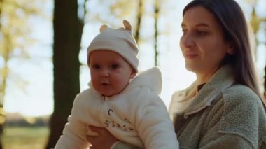 Anne bebeğini kucağında tutarken komik suratlı, kameraya bakan küçük şirin çocuğun portresi. Anne ve bebek ormanda yürüyor. Gündüz vakti. Dışarı çıkın. Ebeveyn, aile