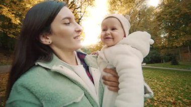 Annesinin ellerinde oturan güzel bir bebeğin vurulması. Anne parkta komik kızıyla yürüyor. Sonbahar geldi. Olumlu duygular. Arka planda sarı ve turuncu yapraklı güzel ağaçlar