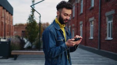Şık saç kesimli, akıllı telefonu elinde tutan ve etrafa bakınan hoş bir genç adamın portresi. Gülümseyen neşeli adam şehirde yürürken iyi haberlerle mesaj okuyor. Gündüz