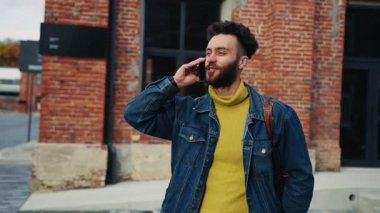 Açık havada akıllı telefondan konuşan yakışıklı, sakallı, beyaz bir adamın görüntüsü. Kendine güveni olan ve şık giyinen konuşkan bir adam. Arkaplanda binalar