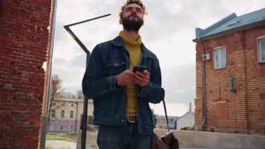 Şehirde tek başına eğlenen gülümseyen genç bir adamın görüntüsü. Sakallı, çekici ve kaygısız bir adamın huzurlu bir ortamda eğlenmesi. Güneş ışığı. Sınavdan sonra mutlu öğrenci. Açık havada.