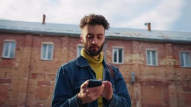 Kendine güvenen, şık saç kesimi olan akıllı telefon navigatörü kullanan, klavyeye yazı yazan ve etrafa bakınan genç adamın yatay görüntüsü. Şehirde kaybolan çekici adamın portresi. Güneş ışığı. Bahar. Dışarıda.