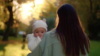 Gün batımında sonbahar parkında yürürken kucağında küçük bir kızı tutan güzel bir kadının arka planı. Gülümseyen anne küçük kızına bakıyor. Açık havada. Şefkat. Annelik. Güneş ışığı