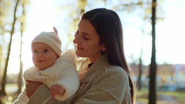 Sevgi dolu bir annenin kucağında sevimli bir bebekle gün batımında güzel bir sonbahar parkında kameranın önünde poz vermesi. Annelik, aile kavramı. Açık havada.