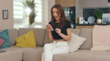 Heyecanlı genç bir kadın telefonda konuşurken koltukta otururken kendini mutlu hissediyor. Güneşli odada gülümsüyor. İnternet sosyal medya bağlantısı. Yavaş çekim