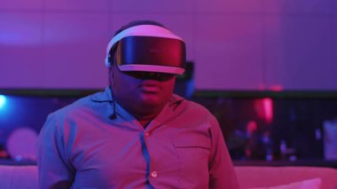 Sanal gerçeklik kulaklığı takmış genç melez bir adam evdeki neon ışıkta kanepede oturuyor. Eğlenceli oyundu. VR gözlükleri sosyal ağ robotu. Yavaş çekim