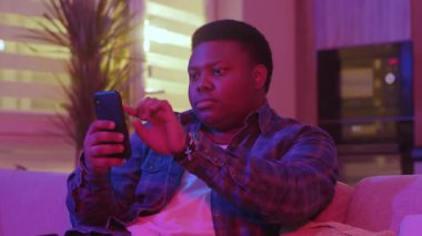 Ciddi bir Afrikalı genç Amerikalı mesaj yazıyor ve akıllı telefon ekranına bakıyor. Kanepede oturup evdeki sosyal ağlarda sohbet etmek. Yavaş çekim