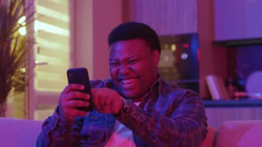Gülümseyen genç Afrikalı Amerikalı adam mesaj yazıyor ve akıllı telefon ekranına bakıyor. Kanepede oturup evdeki sosyal ağlarda sohbet etmek. Yavaş çekim