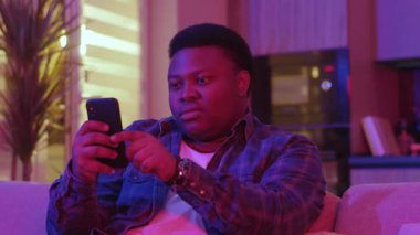 Ciddi bir Afrikalı genç Amerikalı adam mesaj yazıyor ve akıllı telefon ekranına bakıyor. Kanepede oturup evdeki sosyal ağlarda sohbet etmek. Yavaş çekim