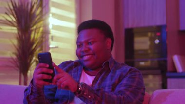 Mutlu gülümseyen genç Afrikalı Amerikalı adam mesaj yazıyor ve akıllı telefon ekranına bakıyor. Kanepede oturup evdeki sosyal ağlarda sohbet etmek. Yavaş çekim