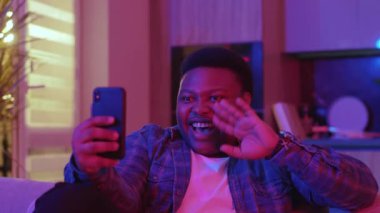 Neşeli, Afro-Amerikalı genç bir adam akıllı telefon ekranına bakarak görüntülü konuşma yapıyor. Kanepede oturup evdeki sosyal ağlarda sohbet etmek. Yavaş çekim