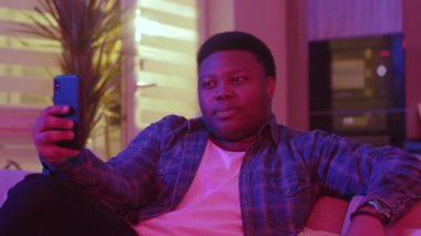 Akıllı telefon ekranına bakarak görüntülü konuşma yapan komik Afrikalı genç bir Amerikalı. Kanepede oturup evdeki sosyal ağlarda sohbet etmek. Yavaş çekim