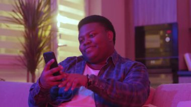 Komik gülümseyen genç Afrikalı Amerikalı adam mesaj yazıyor ve akıllı telefon ekranına bakıyor. Kanepede oturup evdeki sosyal ağlarda sohbet etmek. Yavaş çekim