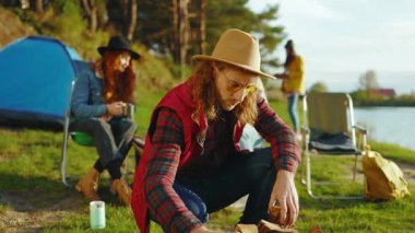 Güneş gözlüklü çekici hippi adam kamp çadırında bir ateş yakıyor. Gülümseyen ve seyahat eden kameralara bakın. Göl kenarındaki ormanda. Kapatın. Yavaş çekim
