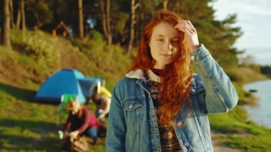 Kızıl saçlı güzel bir kadın kameraya bakarken mutlu oluyor. Arka planda arkadaşları piknik malzemelerini yerleştiriyor. Gün ışığında ormanda gölün yanında dur. Kapatın. Yavaş çekim